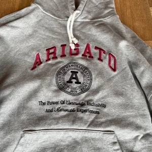 Grå hoodie från Axel Arigato - Säljer en stilren grå hoodie från Axel Arigato med röd text och ett tryck på framsidan. Hoodien har vita snören och en ficka på magen. Säljer för att den jag inte vill ha den längre. Kom gärna med frågor och pris går att diskutera vid snabb affär. 