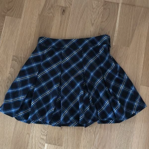 Rutig mini kjol från H&M - Snygg rutig kjol från H&M i blått och svart med vita detaljer. Kjolen har en knappdetalj i midjan och ett klassiskt rutmönster som ger en stilren look. Perfekt för att addera lite mönster till din outfit!