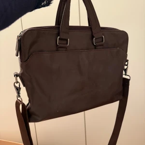 Brun portfölj med axelrem - Snygg brun portfölj med avtagbar axelrem och praktiska handtag. Väskan har en dragkedja upptill och är perfekt för att bära med sig viktiga dokument eller en laptop. Stilren design som passar både till jobbet och skolan. 