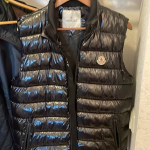 Svart dunväst från Moncler - Snygg svart dunväst från Moncler med glansig finish och dragkedja framtill. Västen har ett klassiskt Moncler-emblem på bröstet och är perfekt för lager-på-lager under kyliga dagar.