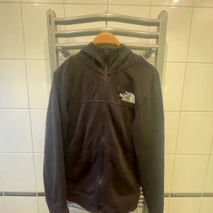 Svart fleecejacka från The North Face - Säljer en svart fleecejacka från The North Face med dragkedja och huva. Jackan har långa ärmar och är perfekt för kyligare dagar. Den är både stilren och funktionell.