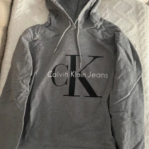  Calvin Klein Jeans - hoodie  - Säljer en stilren croppad grå hoodie från Calvin Klein Jeans med stor logga på framsidan. Tröjan har en klassisk huva med dragsko och långa ärmar. Perfekt för en avslappnad look. Storlek M men funkar också som S! 