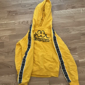 Snygg gul hoodie från kappa med detaljerade ränder på ärmarna - Säljer en gul hoodie från Kappa med svart och vit randig detalj längs ärmarna. Hoodien har en stor ficka framtill och en klassisk Kappa-logga på bröstet. Perfekt för en sportig look! Storleken är 14 år, storlek 162