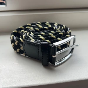 Flätat bälte med svart och gult mönster - Snyggt flätat bälte med ett coolt mönster i svart, gult och vitt. Bältet har en silverfärgad spänne och svarta läderdetaljer vid ändarna. Perfekt för att ge din outfit en unik touch. Vid fler frågor eller bilder, kontakta.