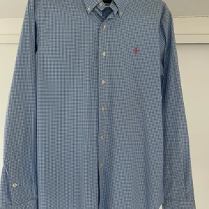 Blå rutig skjorta från Ralph Lauren - Snygg blå rutig skjorta från Ralph Lauren med klassisk krage och knappar framtill. Skjortan har en broderad logotyp på bröstet och långa ärmar. Perfekt för en stilren look.