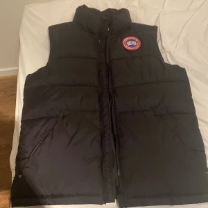 Svart dunväst från Canada Goose - Säljer en svart dunväst från Canada Goose med en broderad logotyp på bröstet. Västen har en dragkedja framtill och är perfekt för kyliga dagar. Den är både stilren och funktionell.