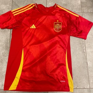 fotbollströja Spanien inga namn - Snygg röd fotbollströja från Adidas med gula detaljer och Spaniens landslagsemblem på bröstet. Tröjan har korta ärmar och tre gula ränder på axlarna. Perfekt för fotbollsfans!