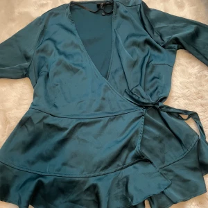 Mörkgrön omlottblus - Elegant mörkgrön omlottblus i satin med v-ringad hals och knytning i midjan. Blusen har långa ärmar och volangdetaljer vid fållen, vilket ger en feminin touch. Förgen glr sig röttvis på sista bilden