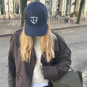 Mörkblå keps med vit logotyp - Snygg mörkblå keps med vit broderad logotyp framtill. Kepsen har ett nätmaterial på baksidan för extra ventilation och en justerbar rem för perfekt passform. 