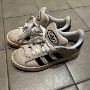 Adidas Campus sneakers - Säljer ett par klassiska Adidas Campus sneakers i beige mocka med svarta ränder. Skorna har en vit sula och snörning framtill. Perfekta för en avslappnad stil. Boxen ingår!