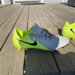 Grå och neongula fotbollsskor från Nike - Säljer ett par grå och neongula fotbollsskor från Nike med snörning. Skorna har en modern design med en iögonfallande färgkombination och är perfekta för fotbollsplanen. Är använda på konstgräs så dom har lite fläckar under skon dom funkar på både gräs och konstgräs storlek 42,5 pris kan diskuteras vid snabb affär