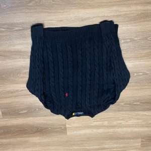 Mörk blå  stickad tröja från Ralph Lauren - Snygg svart stickad tröja från Ralph Lauren med kabelstickat mönster och röd broderad logga på bröstet. Perfekt för kyligare dagar med sin klassiska design och ribbade kanter.