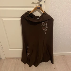 Brun cape från Vasfi Collection - Säljer en elegant brun kjol från Vasfi Collection med vackra broderade blommor i vitt och orange. Perfekt för att lägga till en touch av stil till din outfit. Kjolen har en mjuk och bekväm passform.