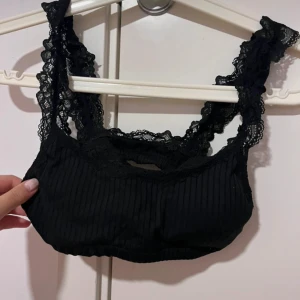 Svart ribbad spets linnetopp med spetsdetaljer - Säljer en svart ribbad linnetopp med breda axelband i spets. Toppen har en croppad passform och är perfekt för lager-på-lager eller som den är. Supersnygg och bekväm med fina spetsdetaljer längs kanterna. Kan användas som BH och topp!