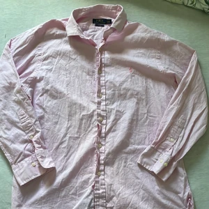skjorta från Polo Ralph Lauren - Säljer en ljusrosa långärmad skjorta från Polo Ralph Lauren i slim fit-modell. Skjortan har klassisk krage, knappar framtill och diskret broderad logga på bröstet. Perfekt för dig som gillar stilrena plagg med en touch av färg. 