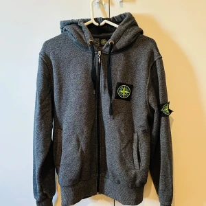 Grå hoodie från Stone Island - Säljer en grå hoodie från Stone Island med dragkedja och huva. Tröjan har två fickor framtill och klassiska Stone Island-loggan på bröstet och ärmen. Perfekt för dig som gillar streetwear och vill ha något stilrent.