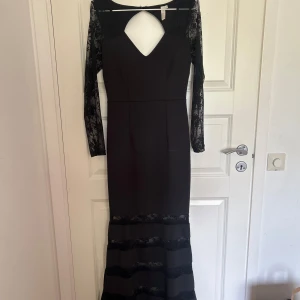 Svart långklänning med spetsärmar och cut-out - Elegant svart långklänning med långa spetsärmar och v-ringning framtill. Klänningen har en snygg cut-out detalj vid bröstet och spetsdetaljer nertill. Perfekt för festliga tillfällen. Klänningen är i storlek 36 men passar lika bra som 38 då den är stretchig.