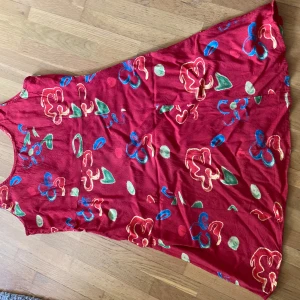 Röd blommig klänning från Vero Moda - Säljer en röd klänning från Vero Moda med färgglatt blommönster i grönt, blått och gult. Klänningen är ärmlös och har en ledig passform, perfekt för varma dagar. Den är tillverkad i ett mjukt och lätt material.