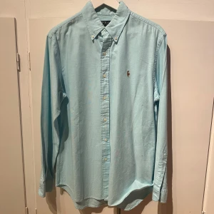 Ljusblå skjorta från Polo Ralph Lauren - Snygg ljusblå skjorta från Polo Ralph Lauren med klassisk button-down krage och broderad logga på bröstet. Slim fit-modell i stretchig bomull, perfekt för en clean och stilren look. Knappar framtill och långa ärmar.