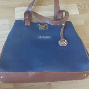 Blå och brun handväska från Michael Kors - Snygg handväska från Michael Kors i blått och brunt skinn. Väskan har guldfärgade detaljer, MK-logga och dragkedja på baksidan. Perfekt storlek för vardagens alla prylar och har två rejäla handtag. Obs ej äkta!