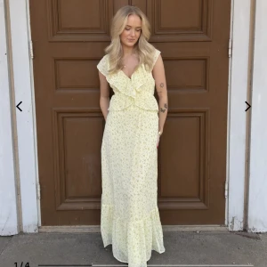 Ljusgul långklänning med volang och blommigt mönster - Söt långklänning i ljusgul färg med små blommor, djup v-ringning och volangdetaljer framtill. Klänningen har öppen rygg med knyt och är ärmlös. Perfekt för sommarens tillfällen.
