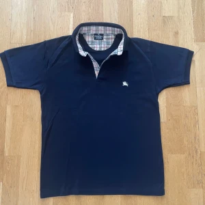 Mörkblå pikétröja från Burberry - Säljer en klassisk mörkblå pikétröja från Burberry med korta ärmar och ikoniskt rutigt tyg på insidan av kragen. Tröjan har en liten broderad logga på bröstet samt en på vänster arm och är perfekt för en stilren look och sommaren! 