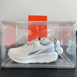 Vita Nike sneakers med silverdetaljer - Säljer ett par fräscha vita Nike sneakers med silverfärgad swoosh och svart sula. Skorna har en modern, sportig design med snörning och rund tå. Perfekta för dig som gillar stilrena och bekväma skor.