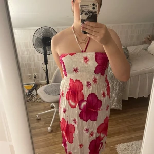 Vit och rosa blommig klänning från Alexander - Säljer en somrig vit klänning från Alexander med stora rosa och röda blommor. Köpt i Grekland och bara använd en gång. Passar mig bra som har M vanligtvis men passar även bra för de som har L eller S då den är smockad. Klänningen är axelbandslös med smock upptill och har en halterneck-knäppning. Tillverkad i 100% rayon, vilket gör den sval och skön.