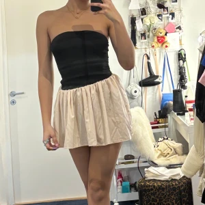 Axelbandslös klänning med svart överdel och beige kjol - Säljer en söt axelbandslös klänning med tight svart överdel och fluffig, ljusbeige kjol. Perfekt för dig som gillar en feminin och trendig stil. Kjolen har lätt volym och överdelen är figurnära.