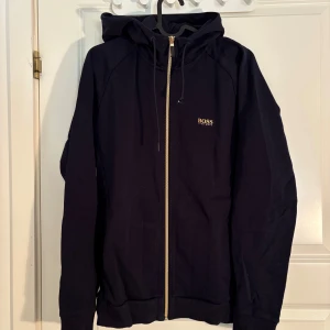 Svart hoodiejacka med dragkedja från BOSS - Svart hoodiejacka från BOSS med guldfärgad dragkedja och huva. Jackan har diskret BOSS-logga på bröstet och ryggen. Perfekt för en avslappnad och stilren look.