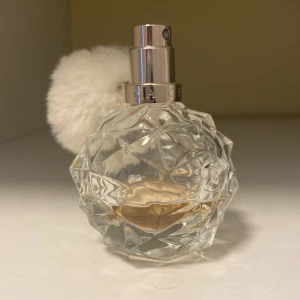 Ari by Ariana Grande Eau de Parfum - Säljer en Ari by Ariana Grande parfym i en kristallformad, transparent flaska med silvrig spraydel och en fluffig vit boll som dekoration. Doften är söt och fruktig, perfekt för dig som gillar unika och lekfulla parfymer.