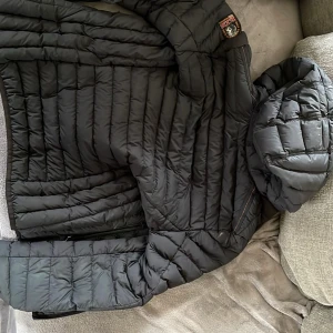 Svart dunjacka från Parajumpers - Säljer en svart dunjacka från Parajumpers med huva och quiltad design. Jackan har lång ärm och är fodrad i ljus färg på insidan. Perfekt för kyliga dagar och har en klassisk look med diskret logga på axeln.