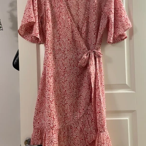 Rosa blommig omlottklänning - Söt rosa omlottklänning med vitt blommönster och volangdetaljer. Klänningen har korta, vida ärmar och knyts i midjan för en smickrande passform. Perfekt för vår och sommar!