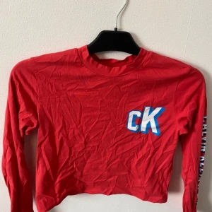 Röd croppad långärmad tröja från Calvin Klein Jeans - Säljer en röd croppad långärmad tröja från Calvin Klein Jeans med CK-logga på bröstet och text längs ena ärmen. Perfekt för en trendig och avslappnad stil. Mjuk bomullskvalitet och rund halsringning.Frakt tillkommer