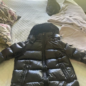 Svart pufferjacka från Moncler - Säljer en svart pufferjacka från Moncler med glansig finish och huva. Jackan har dragkedja framtill, två fickor med dragkedja och en bröstficka med Moncler-logga. Perfekt för kyliga dagar.