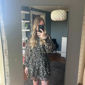 Leopardmönstrad kortklänning med volang - Säljer en snygg kortklänning med leopardmönster och volangdetaljer. Klänningen har långa, transparenta ärmar och markerad midja. Perfekt för dig som vill sticka ut med ett trendigt djurmönster.