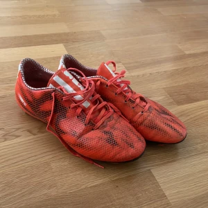 Röda Adidas fotbollsskor med ormskinnsmönster - Säljer ett par röda Adidas fotbollsskor med svart ormskinnsmönster och vita ränder på sidan. Skorna har snörning och dobbar under sulan för bra grepp på planen. Perfekta för dig som vill sticka ut på fotbollsplanen! Vill bara bli av med dom!!!!!