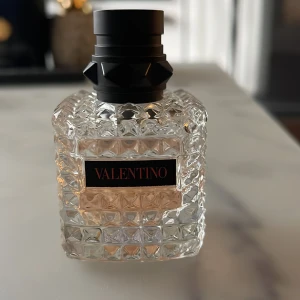 Valentino parfymflaska Donna - Säljer en lyxig parfym från Valentino i en transparent glasflaska med nitar och rutmönster. Flaskan har en svart, robust kork och en elegant etikett med märkesnamnet. Perfekt för dig som gillar exklusiva dofter och snygg design.