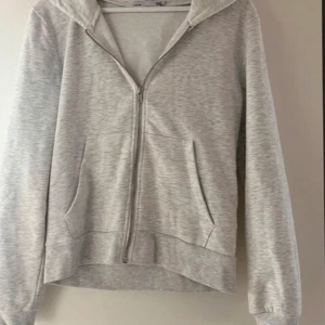 Ljusgrå hoodie med dragkedja - Säljer en enkel och stilren ljusgrå hoodie med dragkedja framtill och två fickor. Perfekt för en avslappnad look. Mjuk och bekväm med långa ärmar och huva.