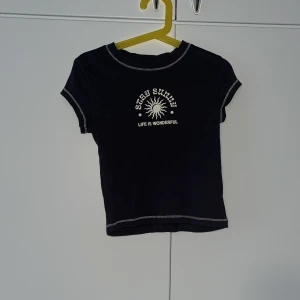 Svart croppad t-shirt med tryck - Svart croppad t-shirt med vit text 'STAY SUNNY LIFE IS WONDERFUL' och soltryck framtill. T-shirten har korta ärmar och kontrastsömmar i vitt. Perfekt för en avslappnad och trendig stil.