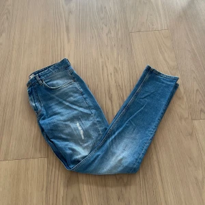 Blå jeans från Dobber - Säljer ett par klassiska blå jeans från Dobber med lätt slitna detaljer framtill. Jeansen har slim passform och raka ben, perfekt för ”grisch” stilen. Fickor fram och bak samt gylf med dragkedja.