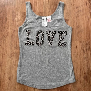 Grå linnetopp / pyjamas med leopardmönstrad text från H&M - Säljer en grå linnetopp från H&M med texten LOVE i leopardmönster framtill. Toppen är ärmlös och har rundad halsringning. Perfekt för en cool och avslappnad stil eller som pyjamas/nattlinne. I fint skick!