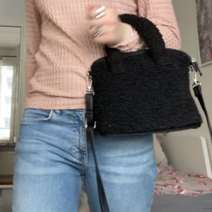 Svart teddy axelväska med handtag - Säljer en svart axelväska i teddy-material med både handtag och lång axelrem. Väskan har en fyrkantig form och är perfekt för att bära det viktigaste. Enkel och trendig design som passar till många outfits.