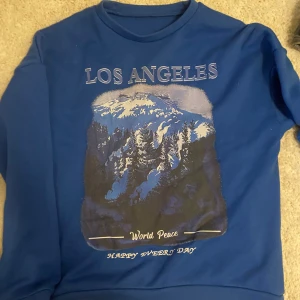 Blå sweatshirt med Los Angeles-tryck - Säljer en blå sweatshirt med tryck av berg och texten 'Los Angeles', 'World Peace' och 'Happy Every Day' på framsidan. 