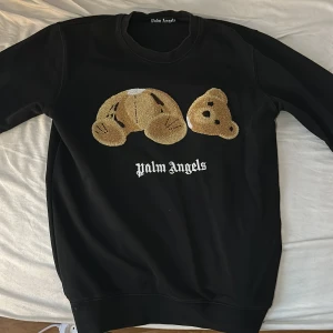 Svart sweatshirt från Palm Angels med nallebrodyr - Svart sweatshirt från Palm Angels med stor broderad nallebjörn på bröstet och vit logotyp under. Tröjan har rund halsringning och ribbade muddar vid ärmslut och nederkant. Perfekt statement-plagg för streetwear.