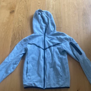 Grå hoodiejacka från Nike - Säljer en grå hoodiejacka från Nike med dragkedja och huva. Jackan har svarta detaljer, två fickor fram och en extra ficka med dragkedja på ärmen. Perfekt för träning eller chill.
