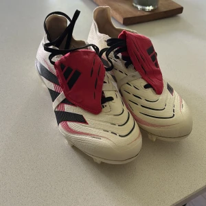 Adidas Predator fotbollsskor i vitt och rött - Säljer ett par Adidas Predator fotbollsskor i vitt med svarta och röda detaljer. Skorna har snörning och klassisk design med täckt snörning och dobbar för spel på gräs. Perfekta för dig som vill ha kontroll på planen. Använda en gång 