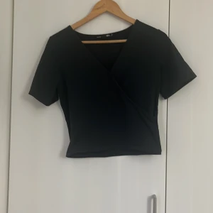 Svart omlottopp från 157 - Svart croppad omlottopp från 157 med korta ärmar och v-ringad hals. Enkel och stilren design som passar till många olika outfits. Perfekt för dig som gillar minimalistiska plagg.