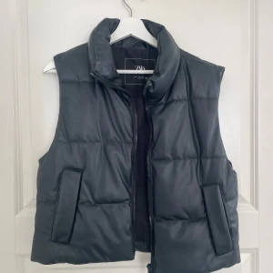 Svart väst från Zara - Säljer en väst från Zara i storlek S. Västen har hög krage, dragkedja framtill och två stora fickor på sidorna. Materialet har en lätt glansig finish och ger en modern look. Använd en sommar