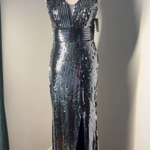 Glittrig långklänning i silver med paljetter - Säljer en maxad långklänning i silver med glittriga paljetter och djup v-ringning. Klänningen har bred midjedetalj, är ärmlös och har en slits framtill. Orginal pris: 2500kr